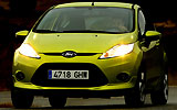Ford Fiesta. Modelo 2009. Imagen. Frontal. Coche en movimiento. Carretera. Luces faros en on. Color verde squeeze.