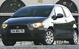 Mitsubishi Colt. Modelo 2009. 3 puertas. Imagen. Exterior. Frontal lateral. Coche parado. Luces faros encendidas. Carretera.